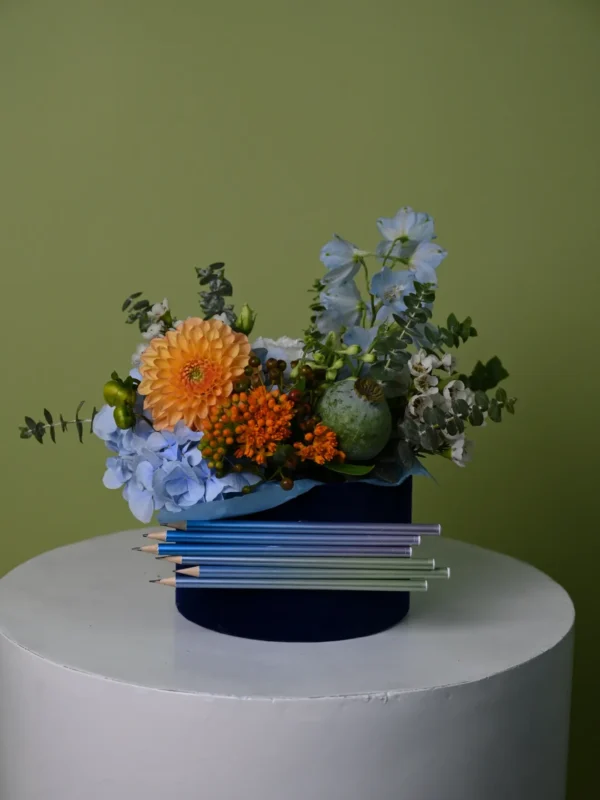 Aranjament în cutie de catifea bleumarin cu hortensie albastră, delphinium, dalie portocalie și creioane